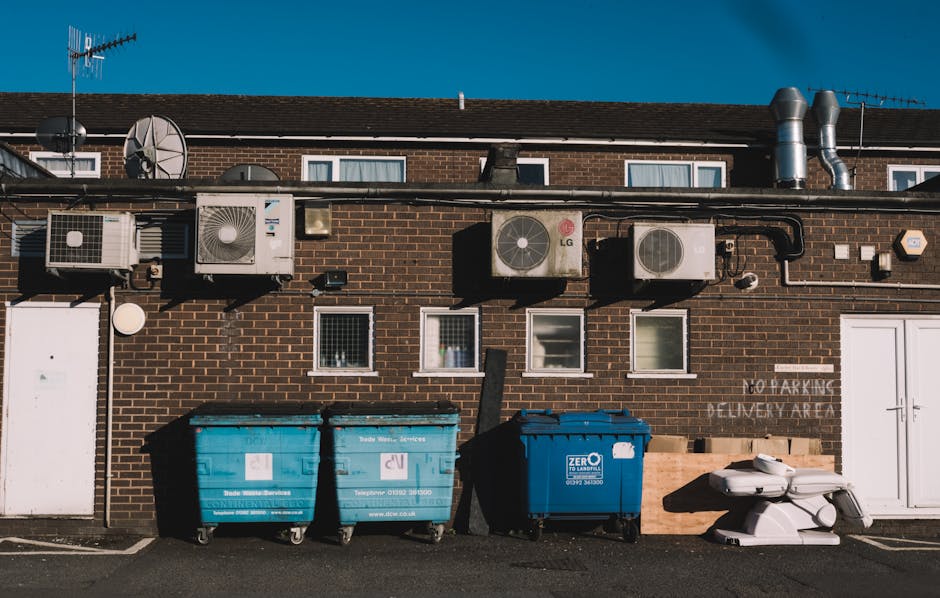 A row of multiple wheeled rubbish bins lined up along a pavement beside a brick wall, with lids in various colours including green, red, blue, and dark green. The bins are made of plastic with texture