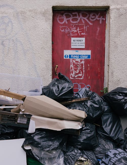 A collection of black rubbish bags, some torn or loosely tied, stacked and piled in front of a red metal door on an exterior wall of a building. The door features multiple graffiti tags in white and p